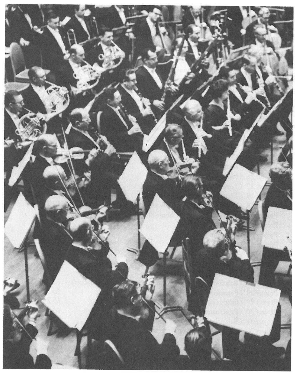Photograph (no printed caption): orchestra players seen from above — horns, clarinets, flutes and violins with music stands.