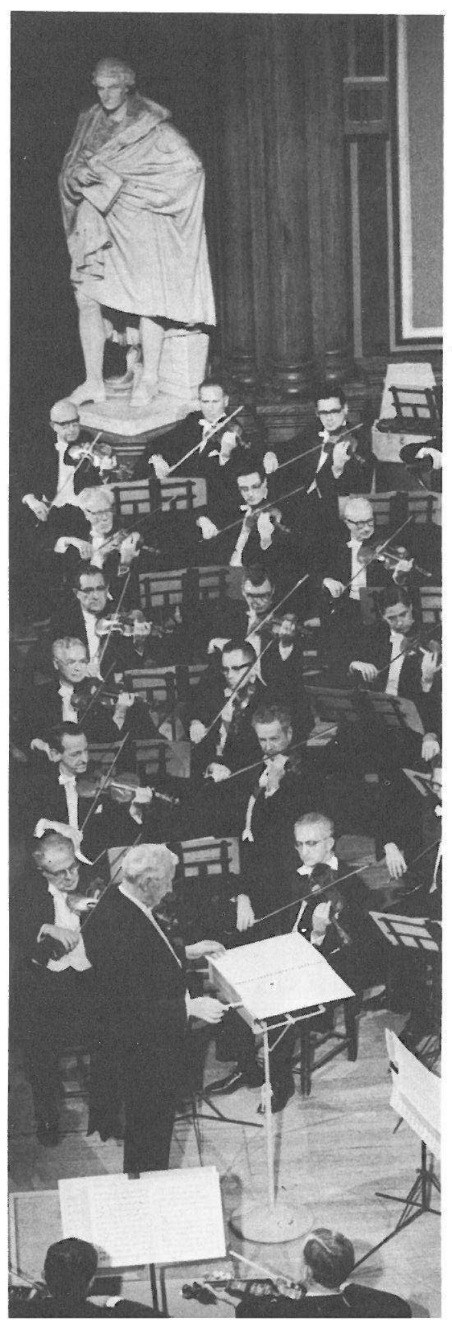 Photograph (no printed caption): the conductor before the violin section, a marble statue on a pedestal behind the players.