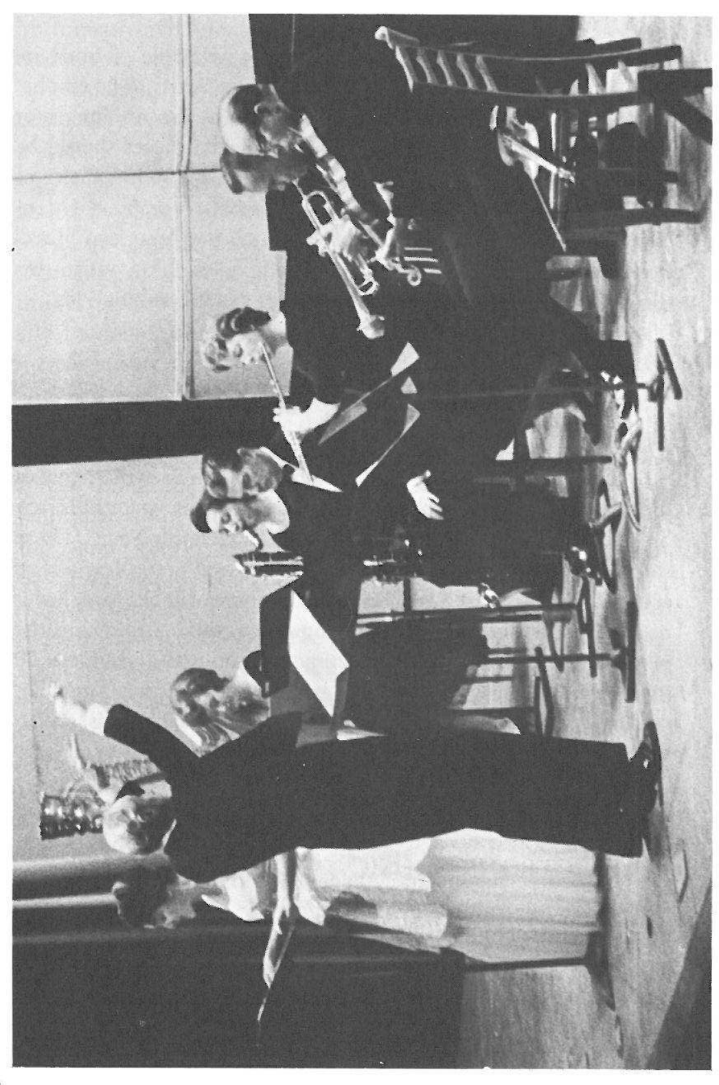Photograph: a small mixed ensemble (trumpet, flute, clarinet and other players at music stands) with the conductor in the foreground; printed rotated 90° on the