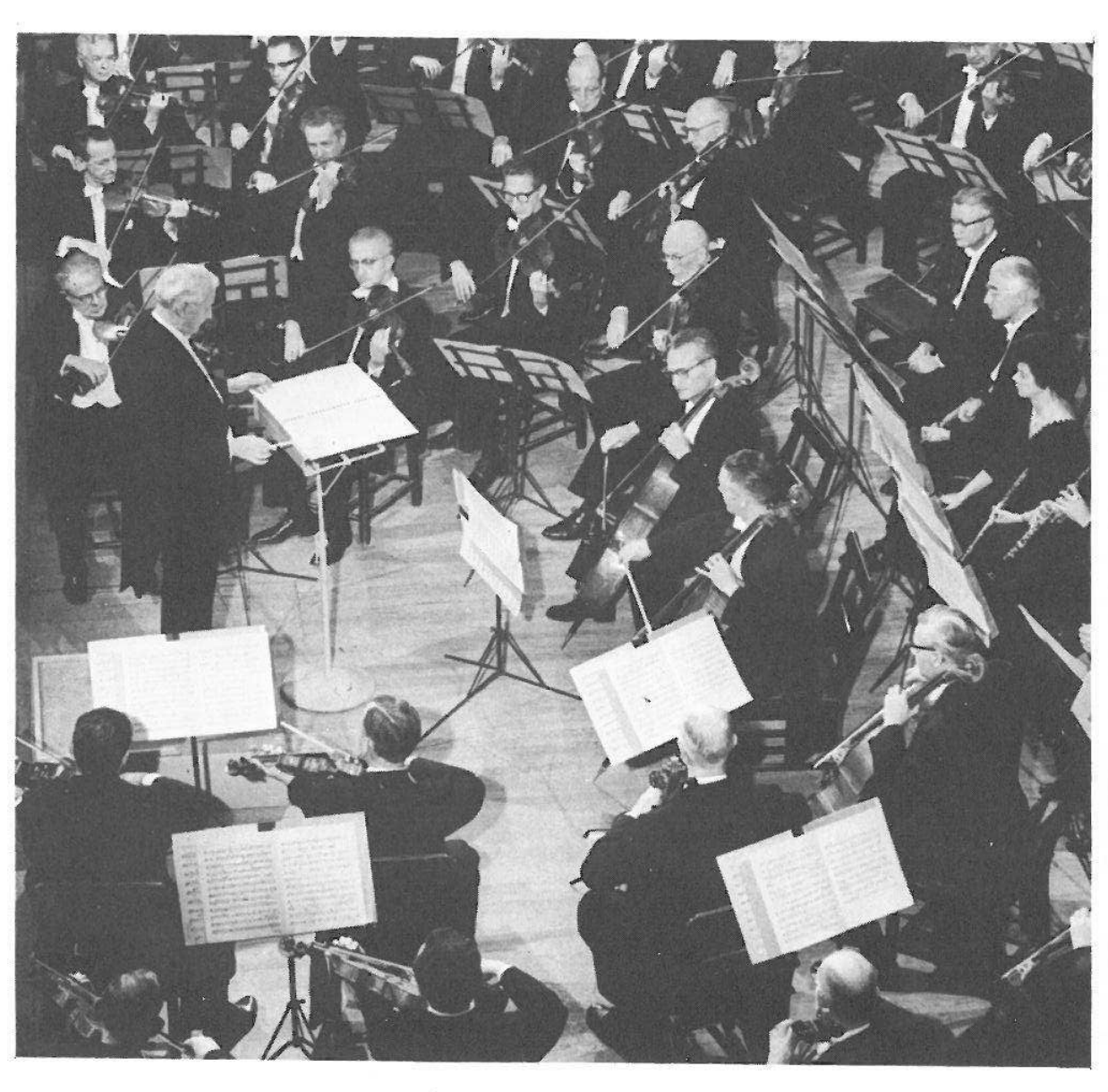 Photograph: an orchestra seen from above, the conductor at his podium among the string players, with woodwinds behind (no caption printed)