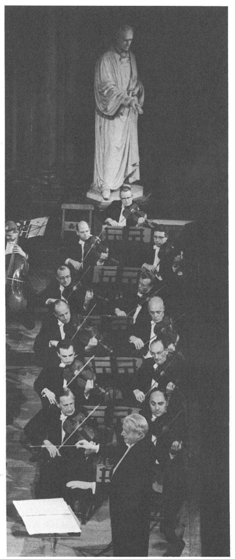Photograph: an orchestra’s string section seen from above, the conductor in the foreground and a statue on a pedestal behind the players (no caption printed)